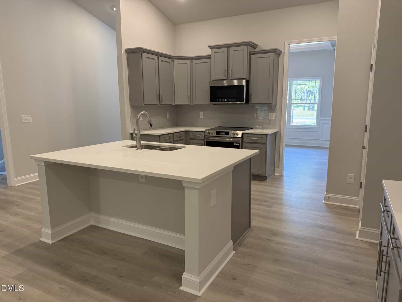 Modern kitchen with gray cabinets, stainless steel appliances, a large island with sink, light wood floors, and bright natural light coming through an adjacent room.