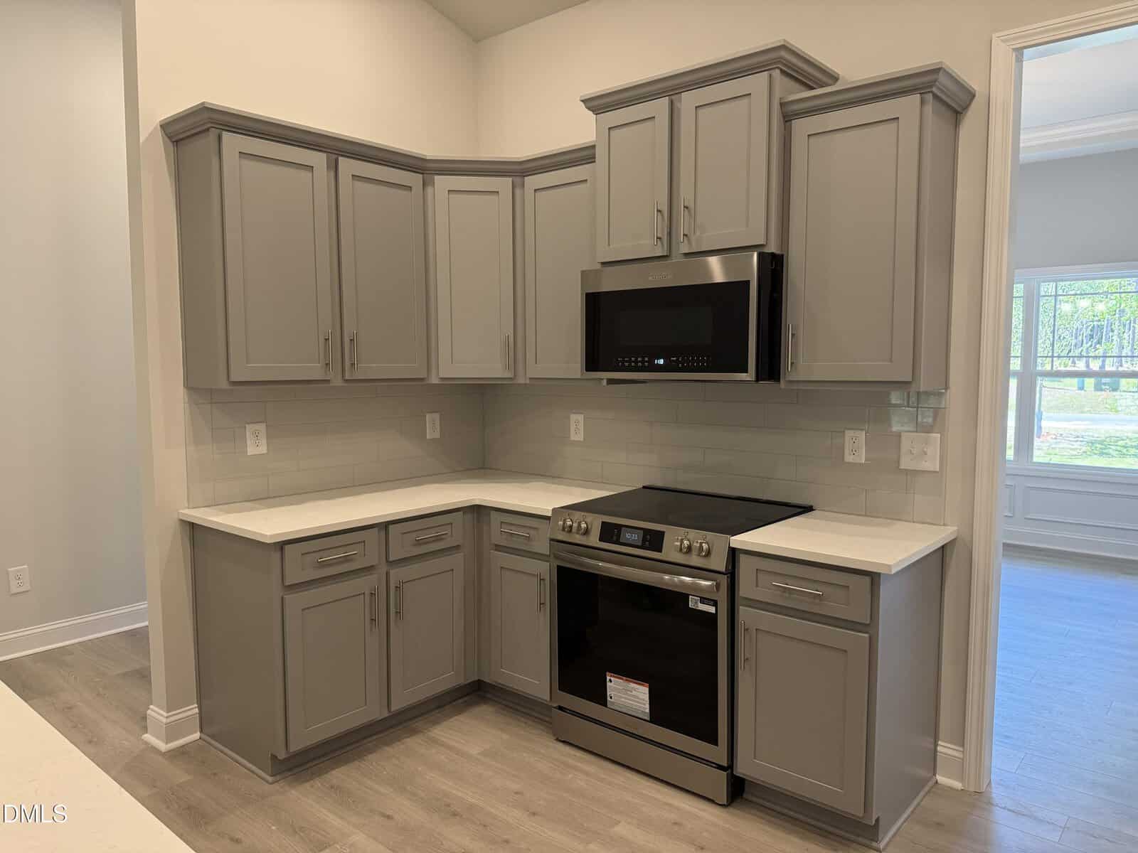 Modern kitchen with gray cabinets, white countertops, stainless steel oven, microwave, and light wood flooring. The walls are light-colored, and there is a window in the adjoining room letting in natural light.