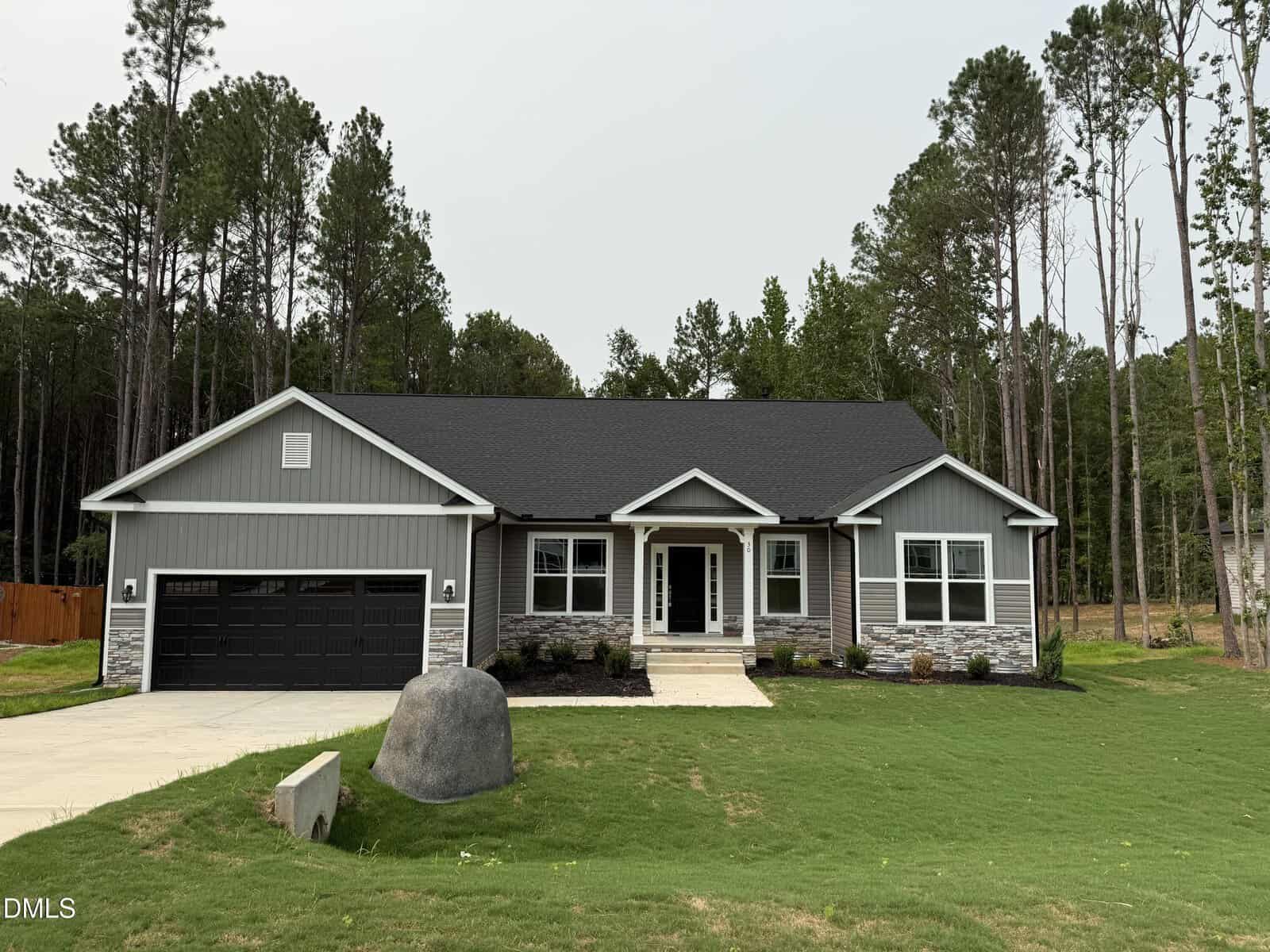 Single-story gray house with black roof, white trim, stone accents, and a black garage door located at 30 Lemon Drop Lane in Franklinton. There's a large rock in the front yard, surrounded by grass and tall trees in the background.