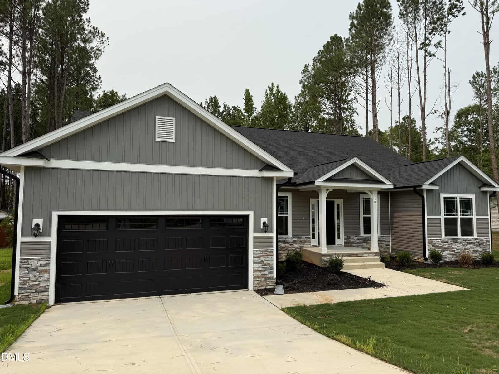 Single-story gray house with black garage doors, white trim, stone accents, a covered front porch, and a neatly landscaped yard, surrounded by tall trees and a concrete driveway.