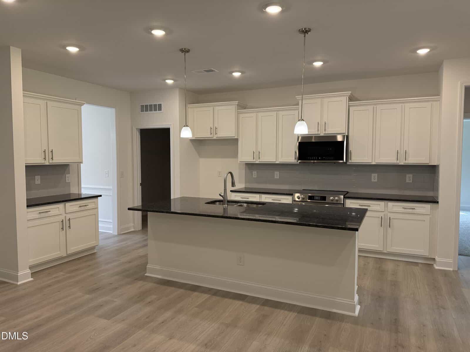 Modern kitchen with white cabinets, stainless steel appliances, black countertops, an island with a sink, light wood flooring, pendant lights, and neutral walls. A doorway leads to another room.