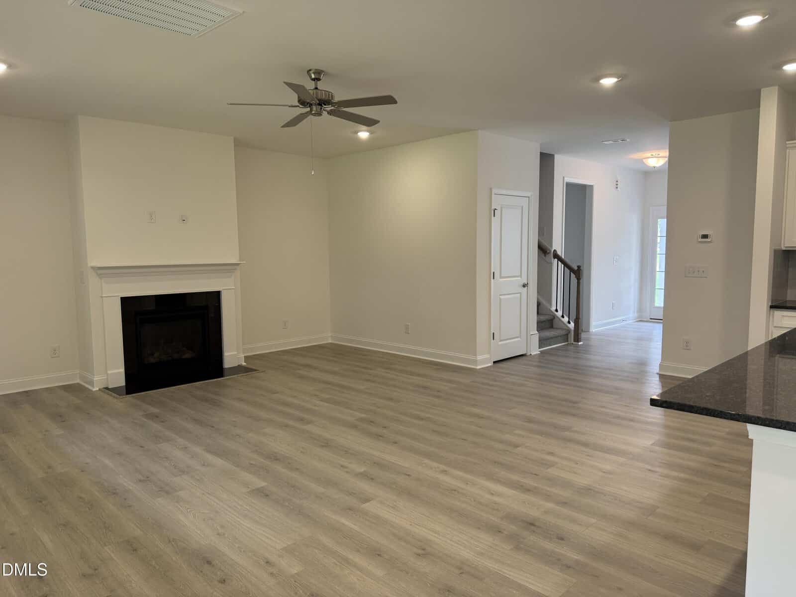 Spacious, empty living area with light wood floors, ceiling fan, fireplace, and stairs leading up. White walls and trim, open to a hallway and kitchen area with dark countertops. Bright natural light fills the space.