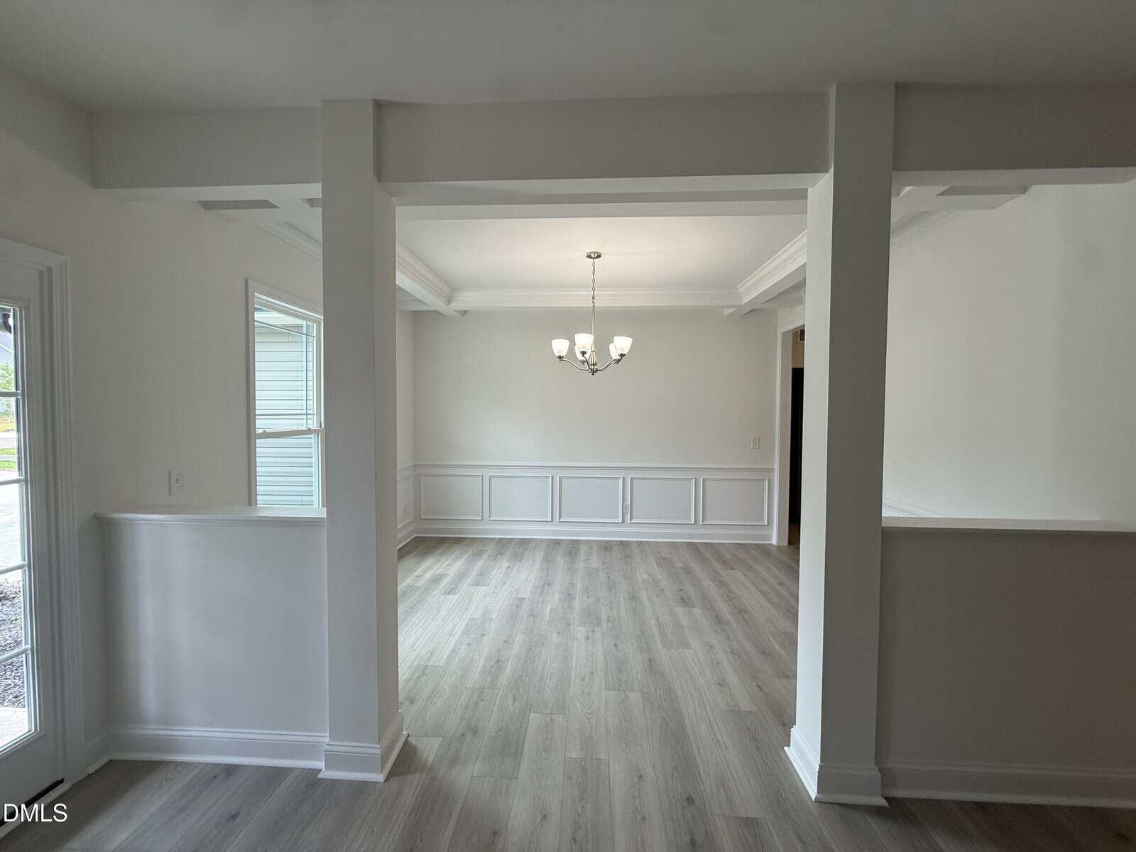 A bright, empty room with light wood floors, white walls, wainscoting, and a ceiling light fixture. The space is divided by partial walls and columns, and sunlight enters through a window and glass door.