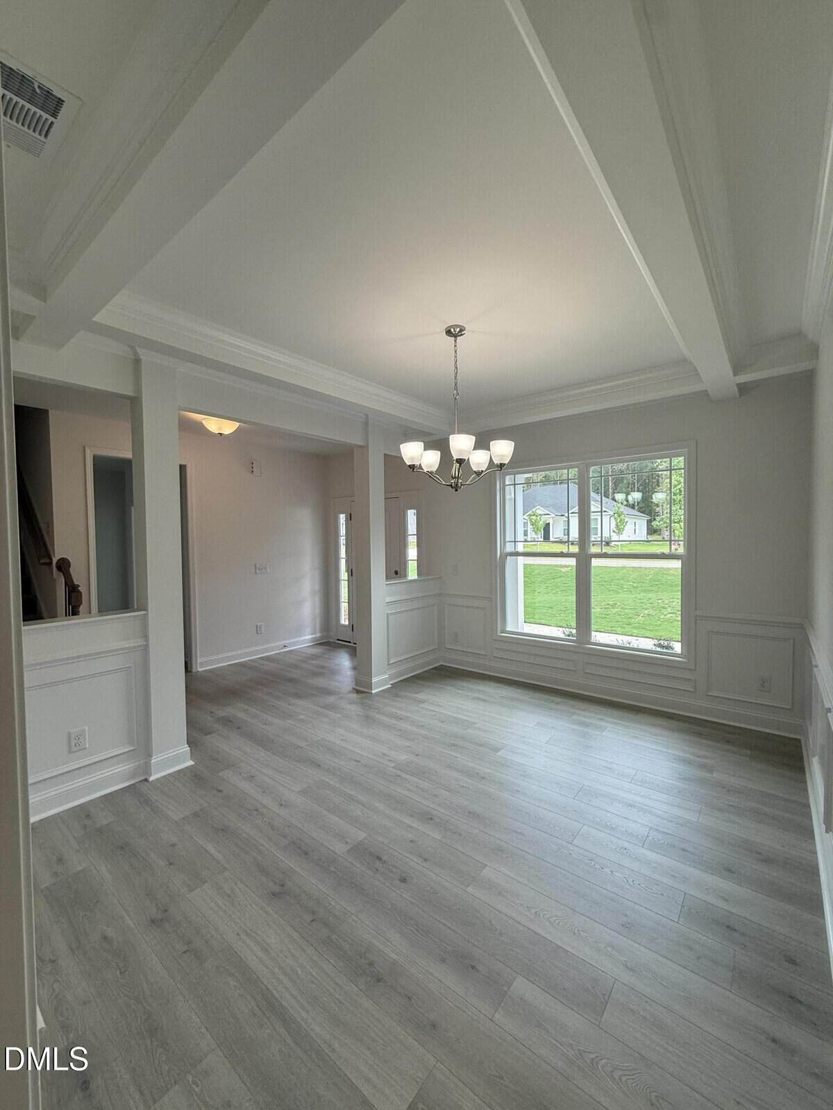 A bright, empty dining room with light wood floors, white walls, a chandelier, large windows showing green grass outside, and decorative ceiling beams.