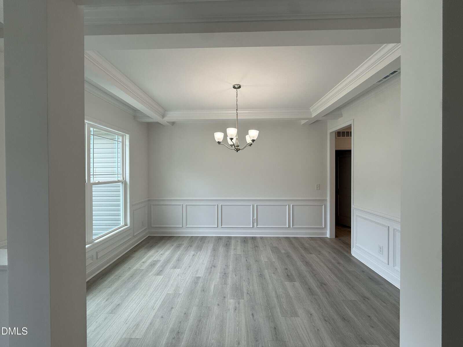 A bright, empty dining room with light wood floors, white walls, large window on the left, wainscoting, tray ceiling, and a chandelier hanging in the center.