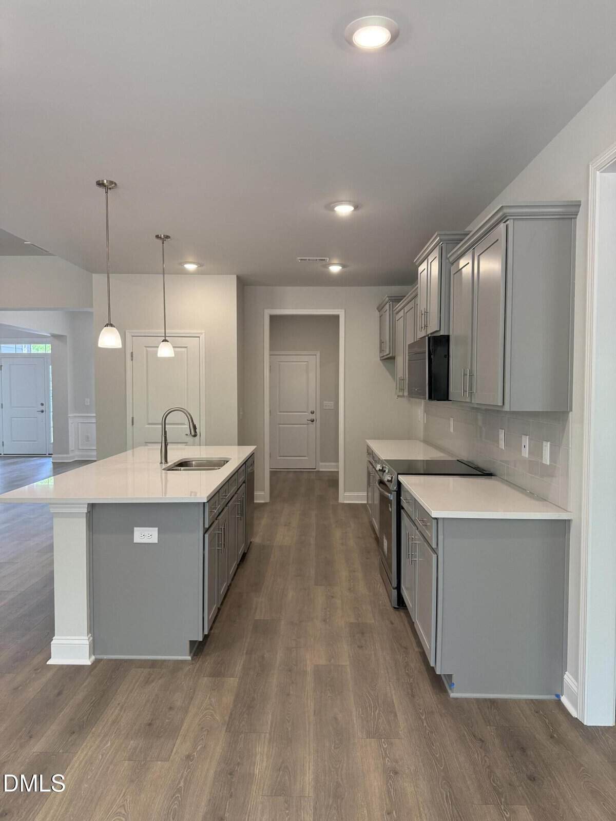 Modern kitchen with light gray cabinets, white countertops, stainless steel appliances, pendant lights above an island with sink, and wood-look flooring. The space is bright with recessed lighting and an open layout.