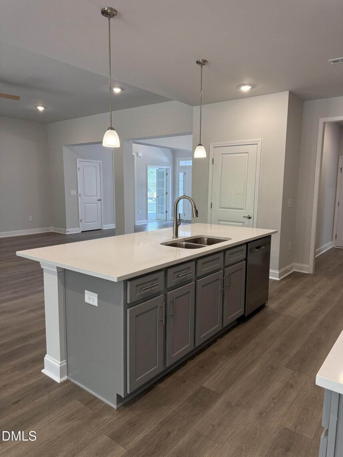 Modern kitchen with a large island featuring gray cabinets, a sink, and pendant lighting. Wood-style flooring and white walls; multiple doorways and ample natural light in the background.
