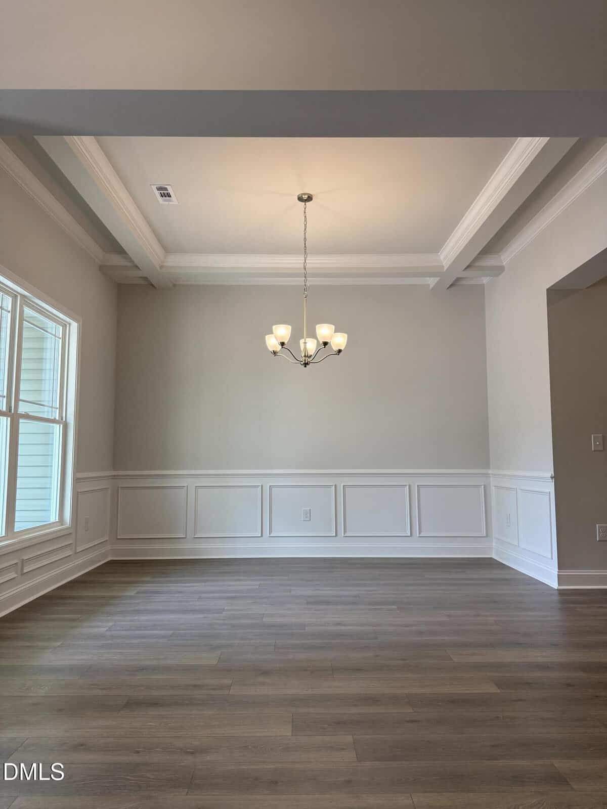 A bright, empty dining room with wood floors, white wainscoting, large windows, and a modern chandelier hanging from a tray ceiling.