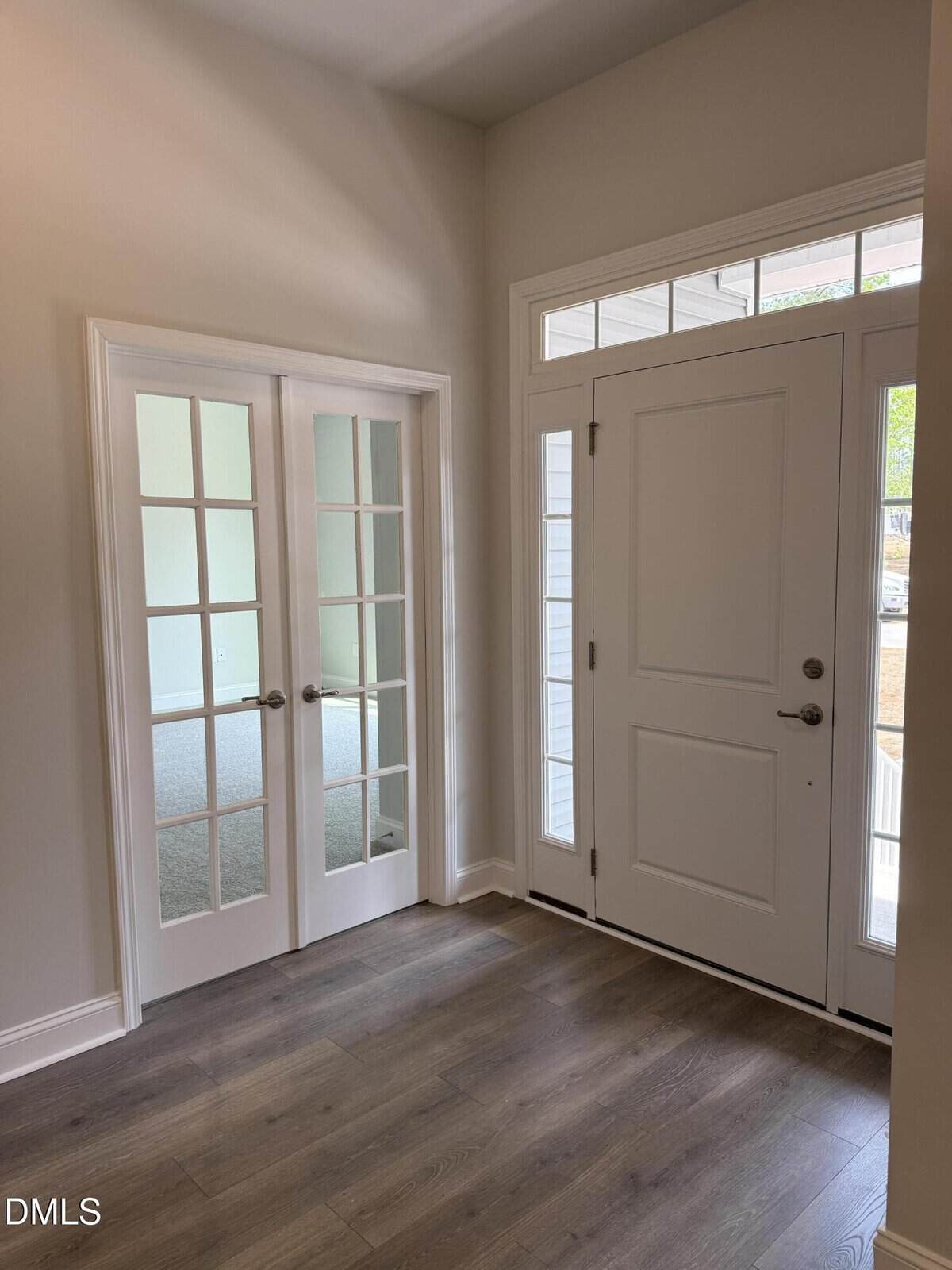 Bright entryway with wood floors, a white front door featuring glass panels, and adjacent double French doors with glass panes, leading into another room. Natural light streams in through windows above the door.