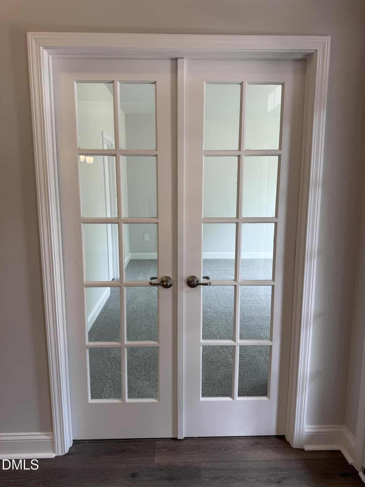 A pair of closed white French doors with glass panes, silver handles, and white trim. The doors lead to a carpeted room with neutral-colored walls. The floor outside the doors is dark wood.