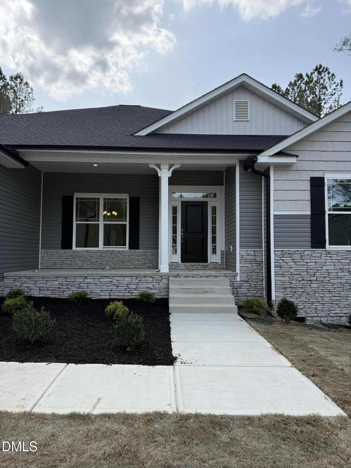 A modern house with gray siding, stone accents, black shutters, and a covered porch. A concrete walkway leads to the black front door. Small shrubs are planted along the front lawn.