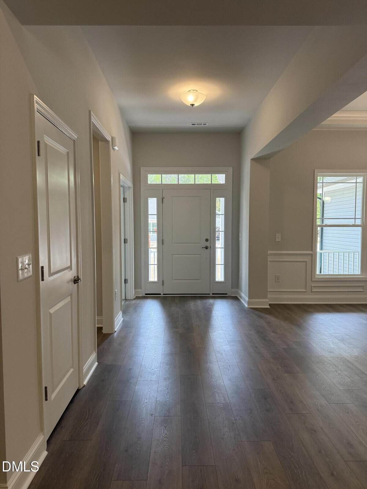 A view of a home's entryway with a white front door, transom windows above it, and adjacent rooms visible. The floors are dark wood, and the walls are painted light beige. Natural light fills the space.