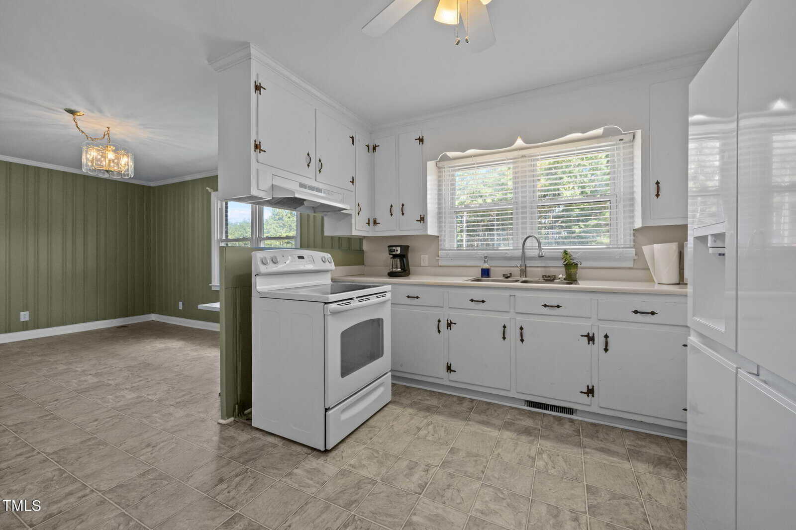 A kitchen with white cabinets, white appliances, and a double sink under a window with blinds. The floor is tiled, and an adjacent dining area with green walls and a chandelier is visible.