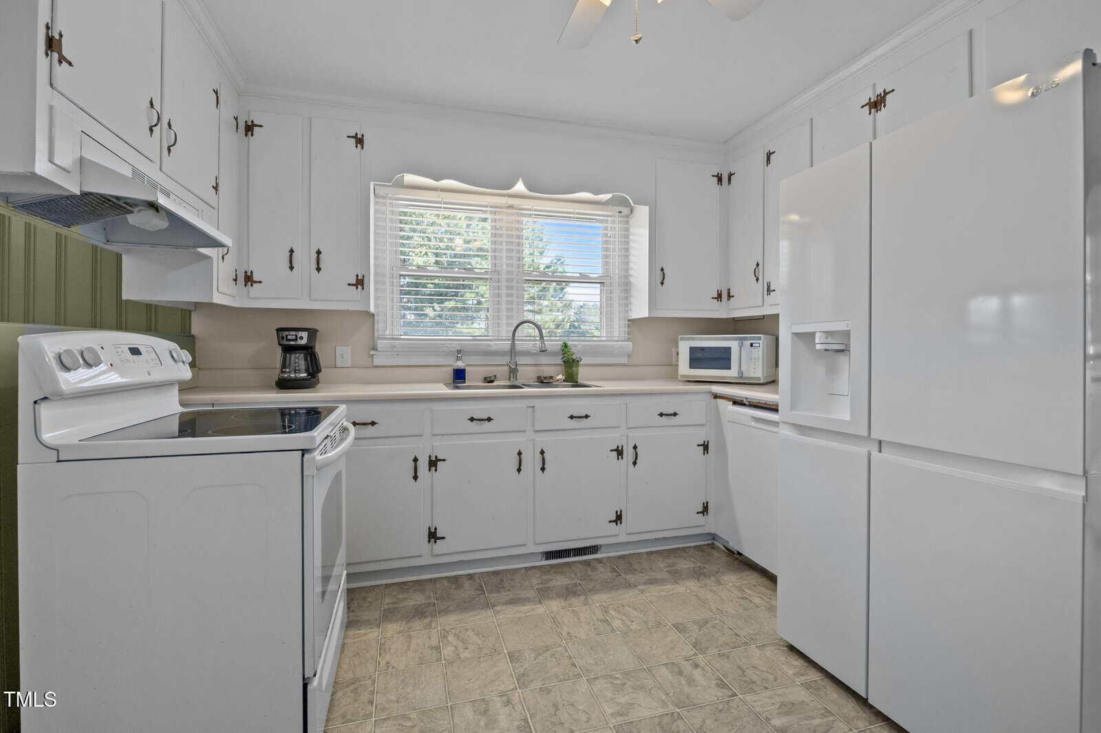 A kitchen with white cabinets, white appliances including a refrigerator, stove, and microwave, a coffee maker, a double sink under a window with blinds, and beige countertops and backsplash.