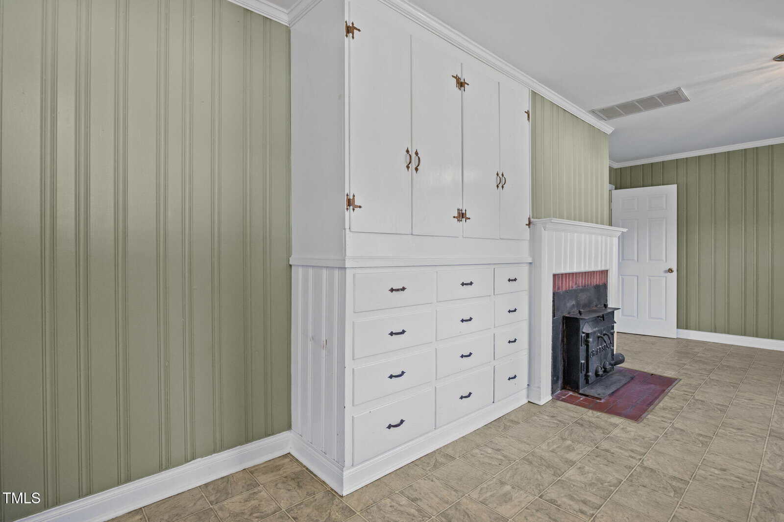 A room with green vertical paneled walls, white built-in cabinets with drawers and cupboards, a small black fireplace with a white mantel, and beige tile flooring. A closed white door is visible in the background.