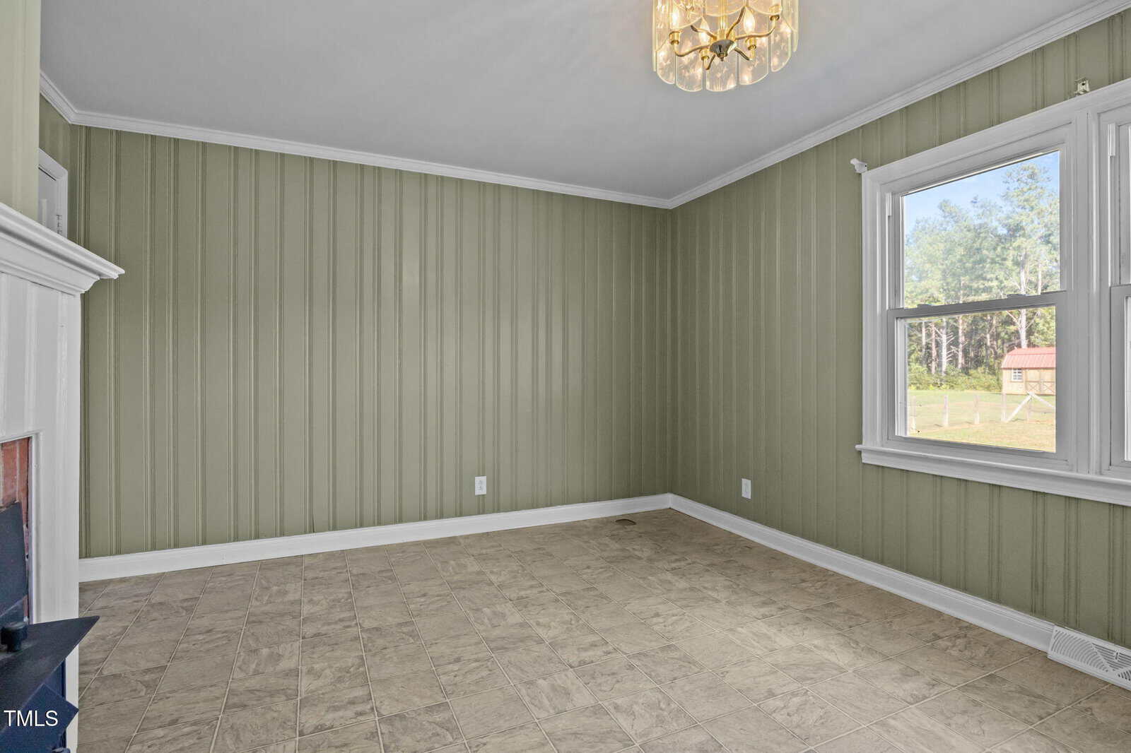 Empty room with light green vertical wood panel walls, beige tile flooring, a large window showing outdoor greenery, a white ceiling, white trim, and a gold chandelier hanging from the ceiling.