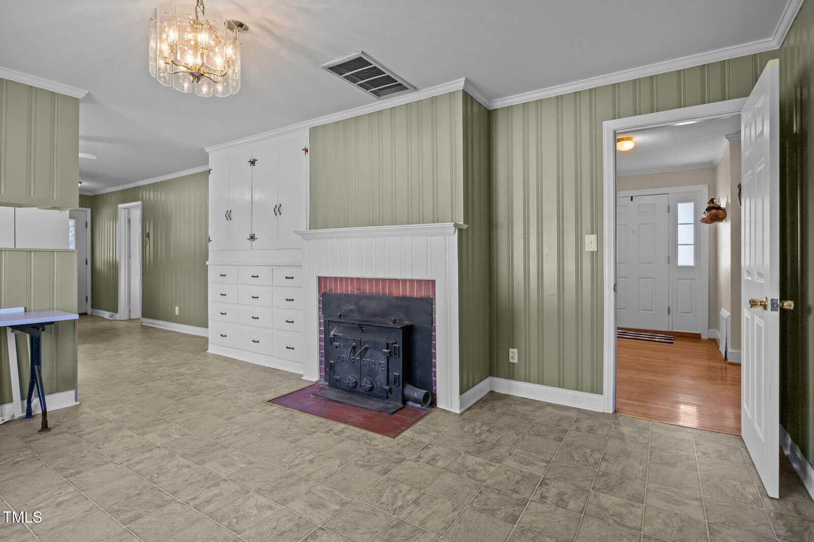 Living room with green paneled walls, white trim, built-in cabinets, and a fireplace with a black cover. Tile flooring, a glass chandelier, and visible entry hallway and doors.