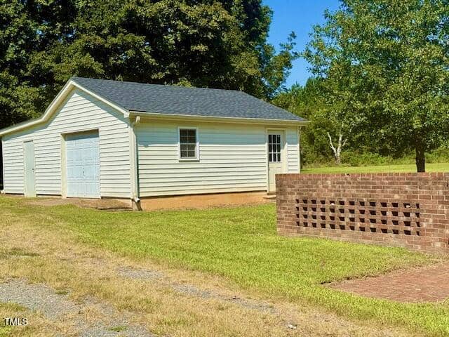 A single-story white garage with a gray roof stands on a grassy lawn beside a small brick wall with decorative openings. Trees and a clear sky are visible in the background.