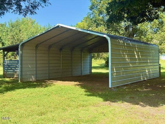 A metal carport with an open front and sides and a covered roof sits on a grassy area, with trees and another small structure visible in the background. Sunlight casts shadows on the ground.