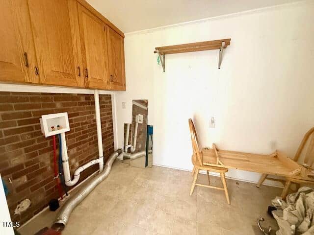 Laundry room with a brick accent wall, wooden cabinets, exposed washer and dryer hookups, a floor mirror, and a small wooden table with two chairs, one of which is tipped over.