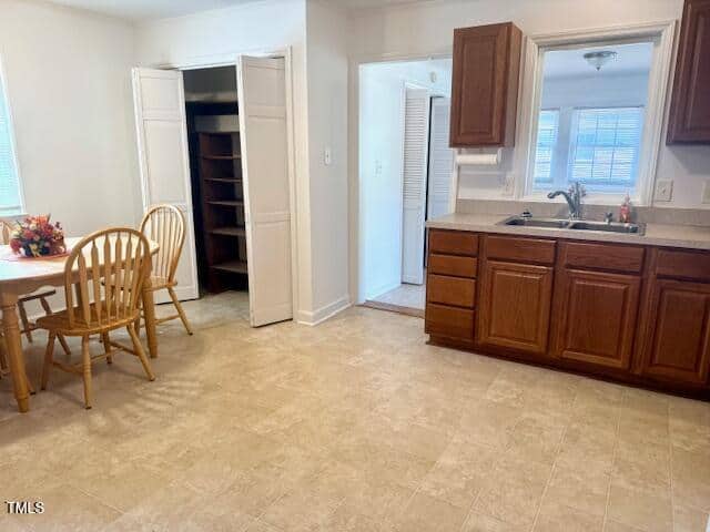 Kitchen with brown cabinets, sink, and beige floor. A wooden dining table with two chairs sits near an open closet with shelves. Natural light comes from windows in the background.