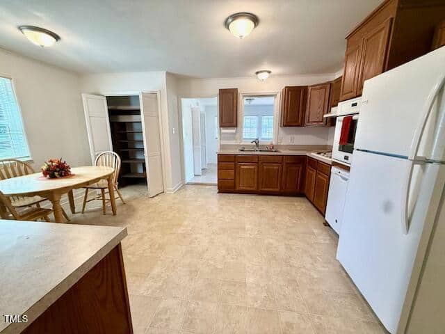 A spacious kitchen with wood cabinets, white appliances, a round dining table with four chairs, tiled flooring, and an open pantry with shelves. Multiple ceiling lights and windows provide ample illumination.