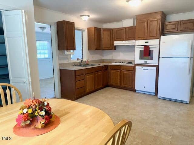 A kitchen with brown cabinets, white appliances including a refrigerator, dishwasher, oven, and range. There is a round wooden dining table with floral centerpiece in the foreground and a pantry with open shelves on the left.