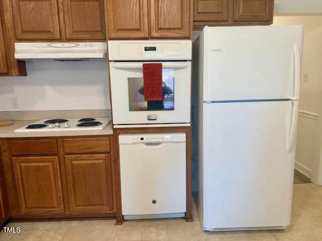 A white refrigerator stands next to a built-in white oven with a red towel hanging on its handle, a white dishwasher below, and a white stove with a range hood, all surrounded by brown wooden cabinets.
