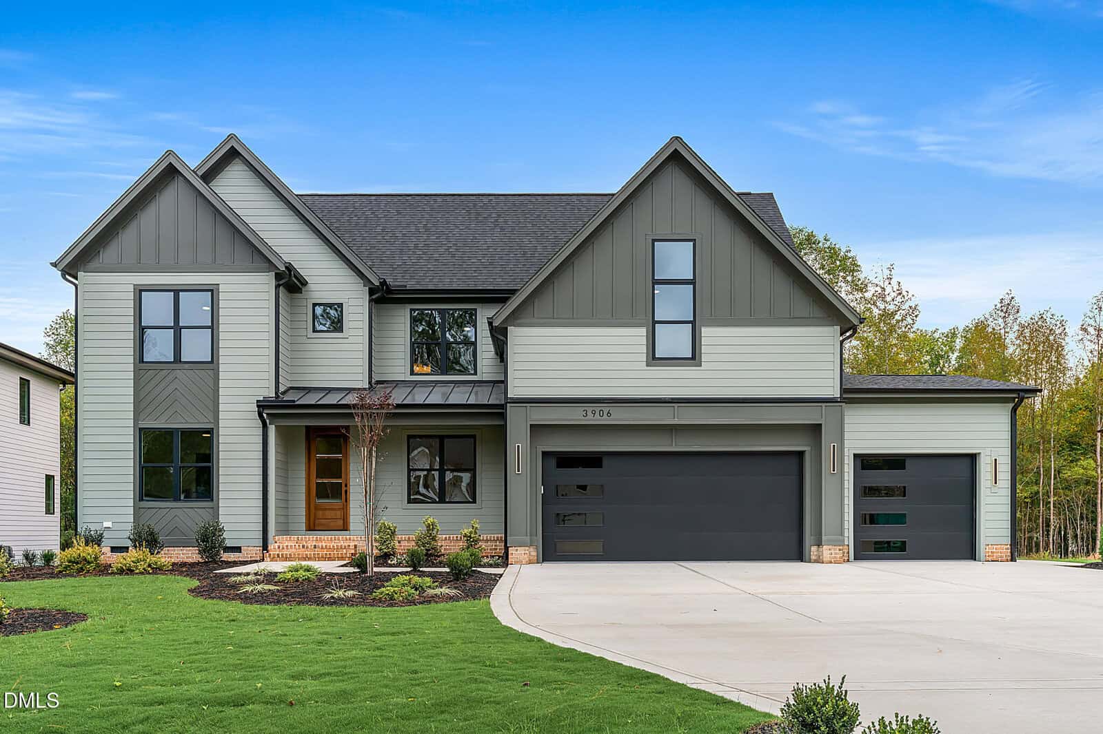 A modern two-story house at 3906 Cedar Knolls Drive, Youngsville, with gray siding, dark trim, large windows, and three garage doors. The front yard has green grass, young trees, and a wide driveway under a clear blue sky.