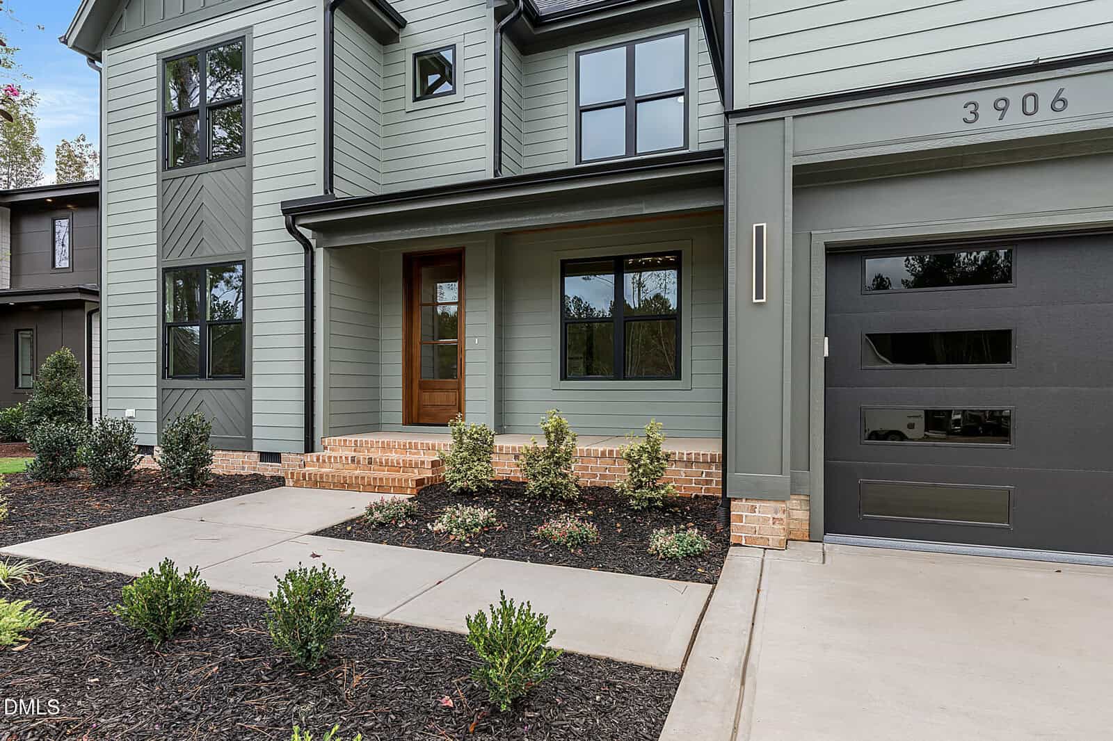 A modern two-story house with light green siding, large black-framed windows, a covered front entry with brick steps, a black garage door, and landscaped bushes along a concrete walkway.