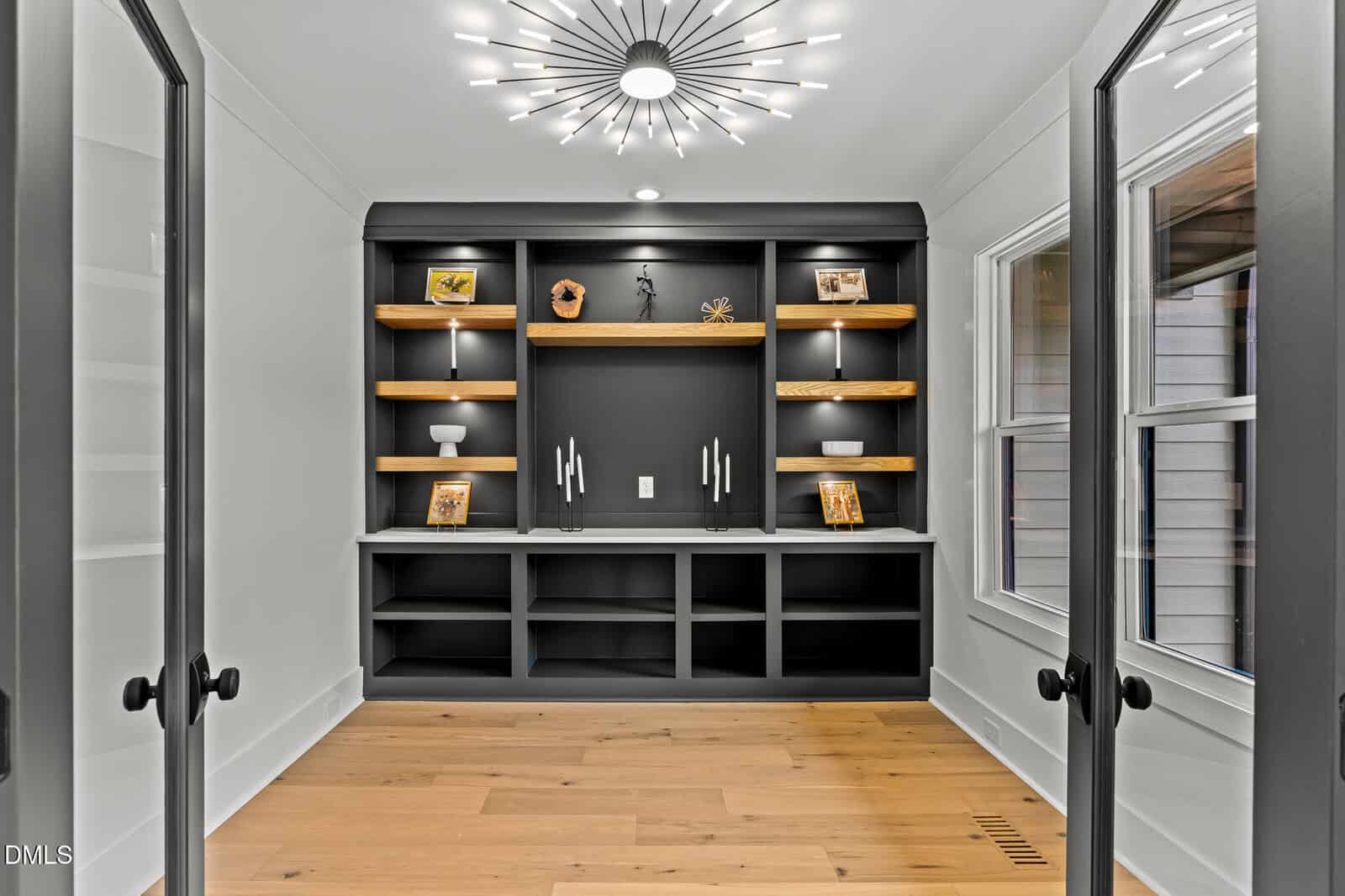 A modern home office features built-in black shelves with wooden accents, decorative items, and framed photos. Double glass doors and large windows let in natural light. A contemporary chandelier hangs from the ceiling.