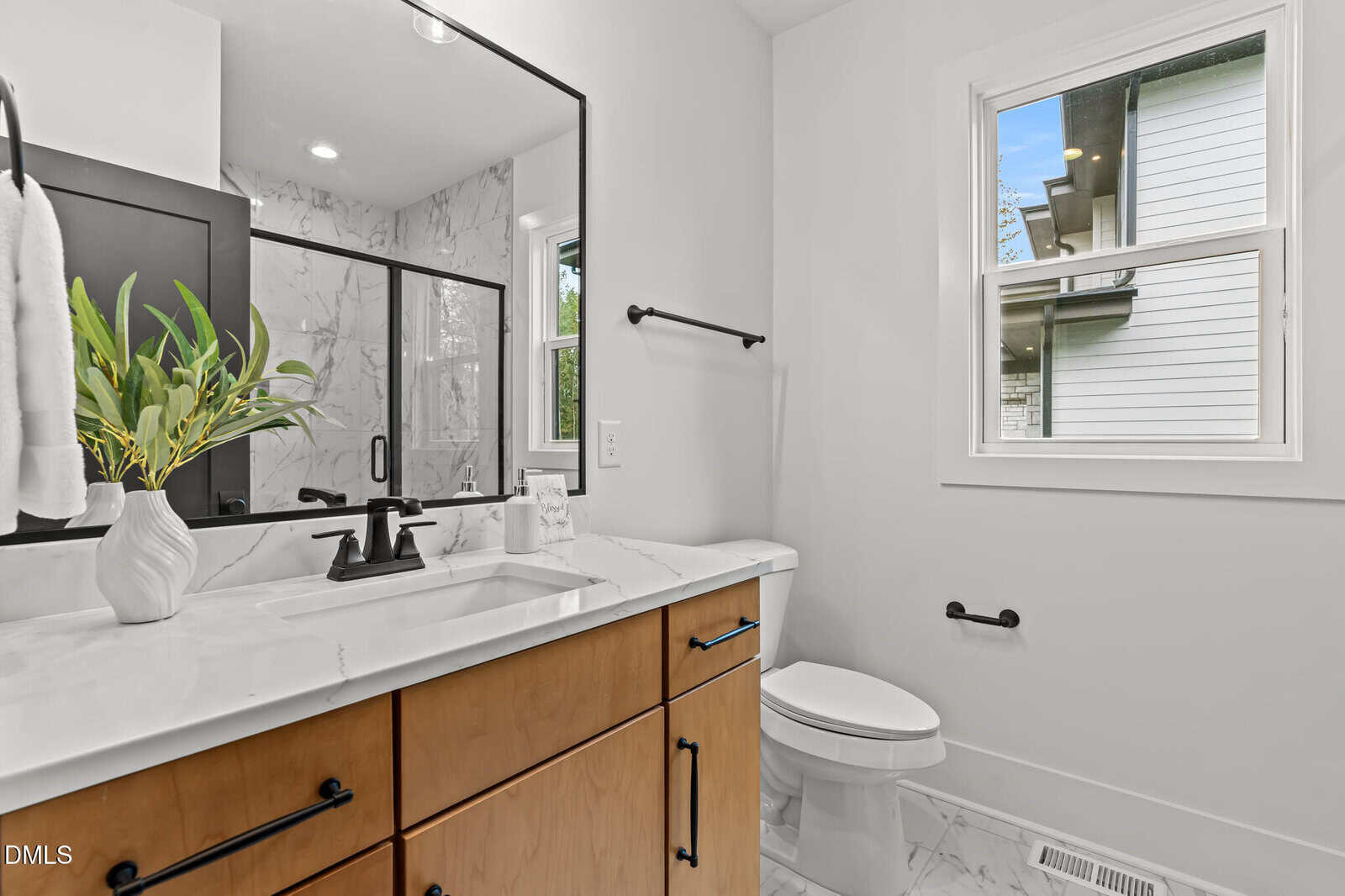 Modern bathroom with a wood vanity, marble countertop, black fixtures, a plant in a white vase, glass shower enclosure, toilet, and a window with a view of a neighboring house. White walls and tile floor.