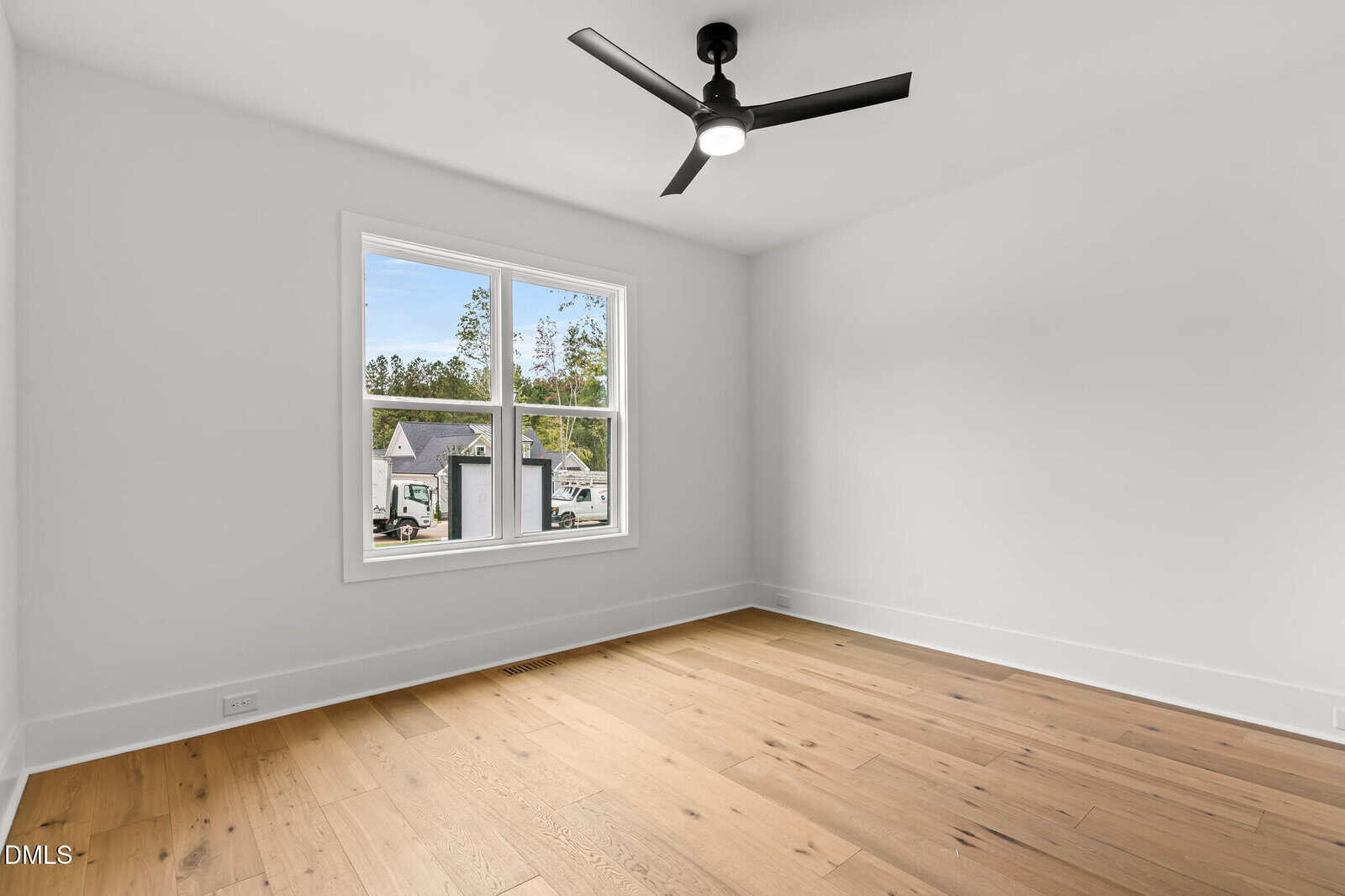 A bright, empty room with light wood flooring, white walls, a large window showing houses and trees outside, and a black ceiling fan with a light in the center of the ceiling.