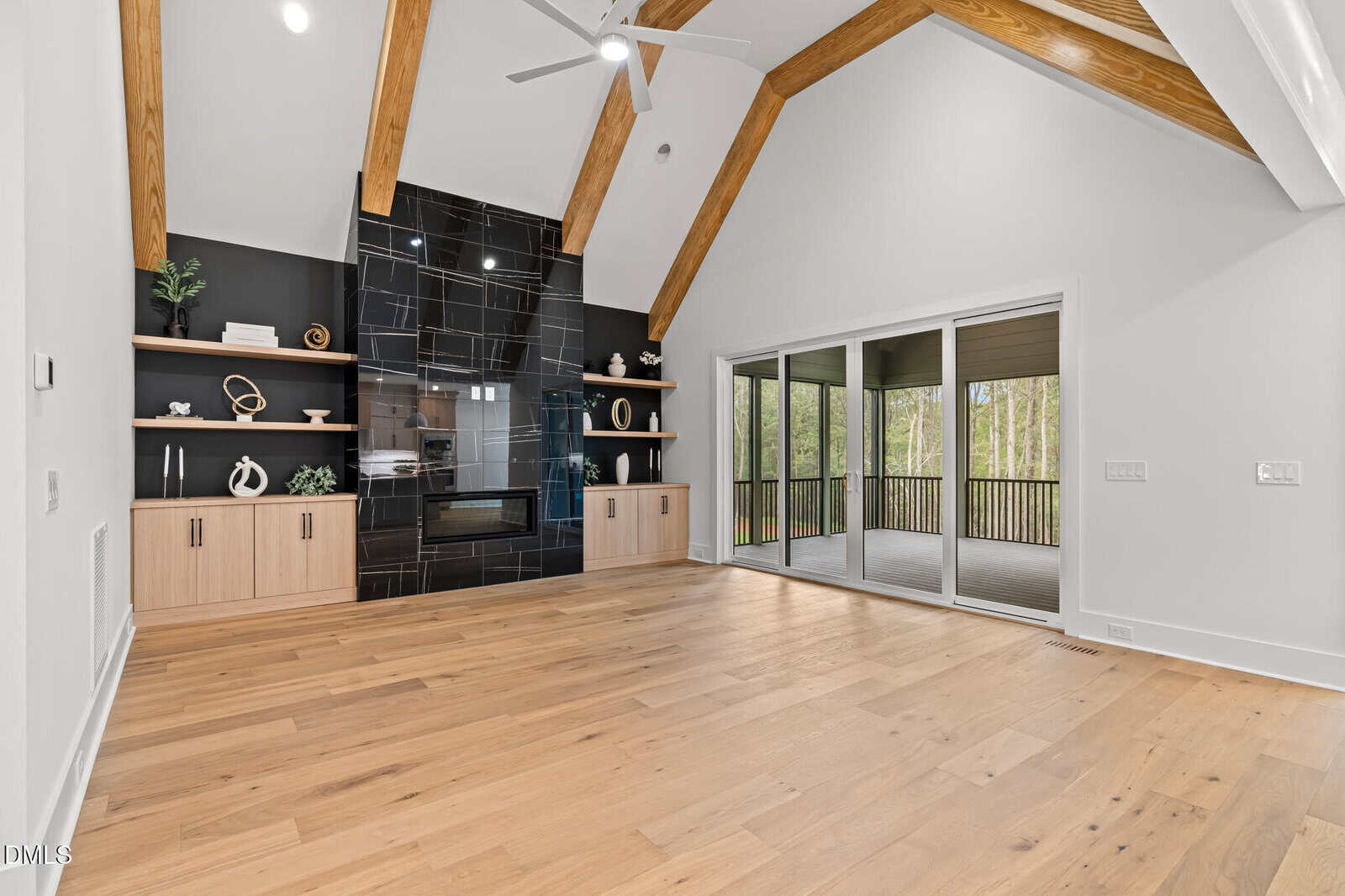 A modern living room with light wood floors, vaulted ceiling with exposed wooden beams, black tile fireplace, built-in shelves and cabinets, and large sliding glass doors leading to a screened porch.