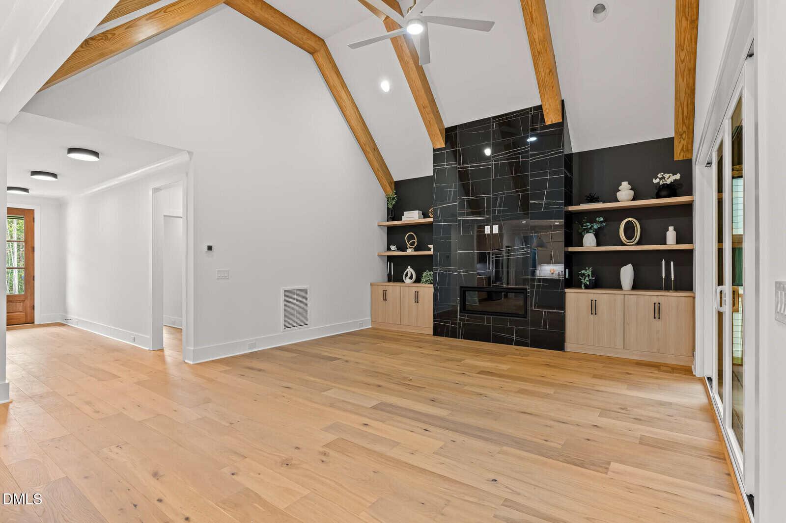 A spacious living room with light wood flooring, vaulted ceiling with exposed wood beams, and a black tiled fireplace flanked by built-in shelves and cabinets. Sliding glass doors are on the right wall.
