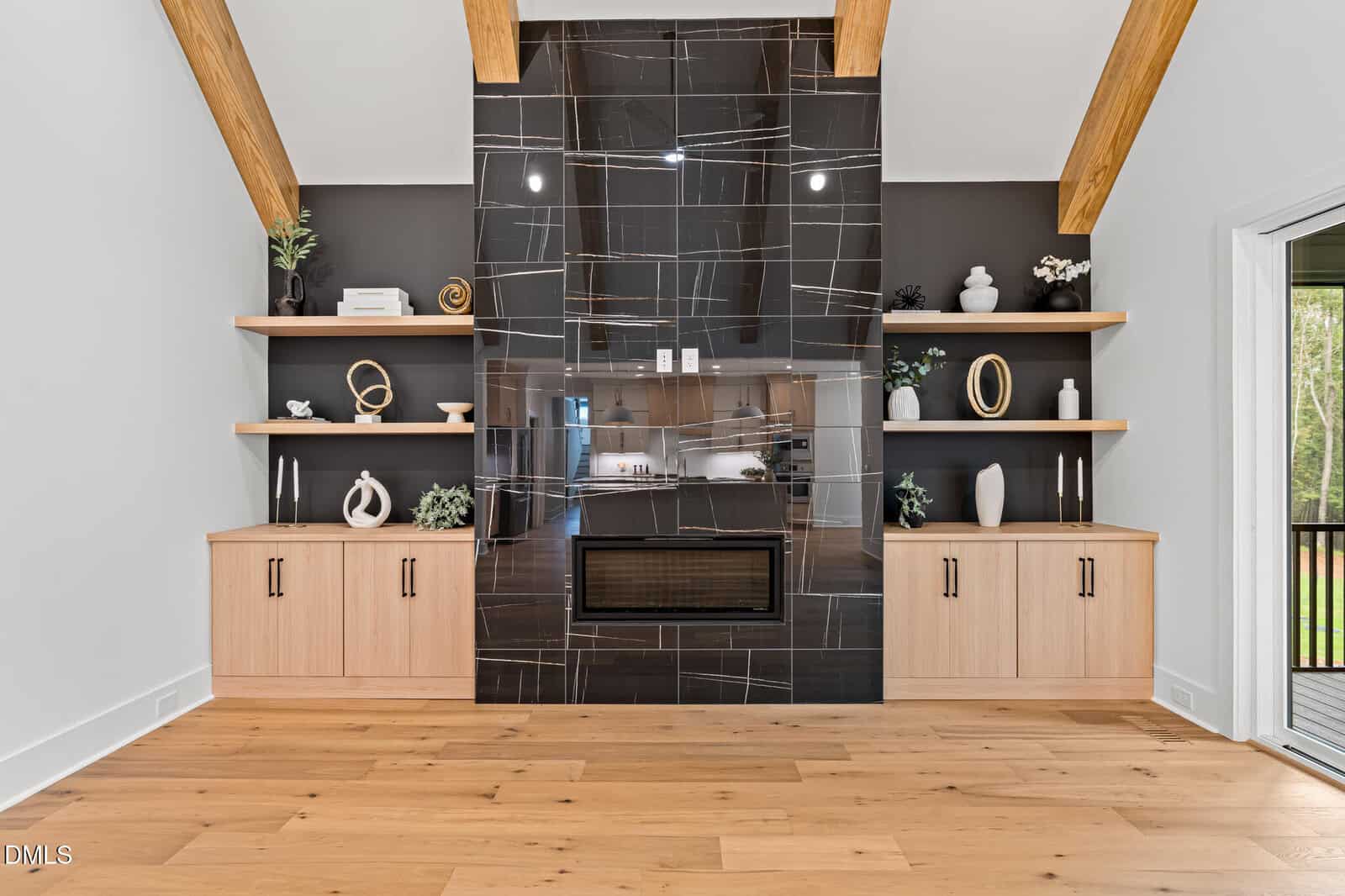 A modern living room with light wood floors, a large black tile fireplace with white lines, open wood shelves with decorative items, and light wood cabinets below, flanked by exposed wooden ceiling beams.