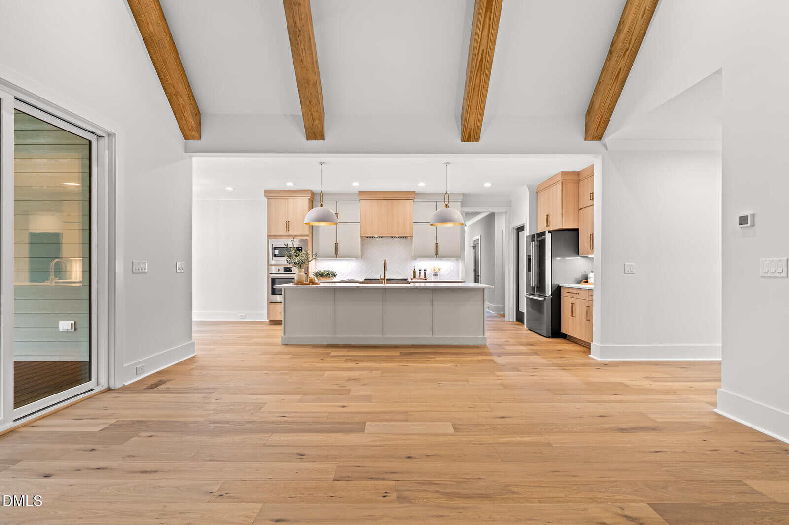 Spacious modern kitchen with light wood floors, exposed ceiling beams, center island with pendant lights, light wood cabinetry, stainless steel appliances, and large glass door leading to another room.