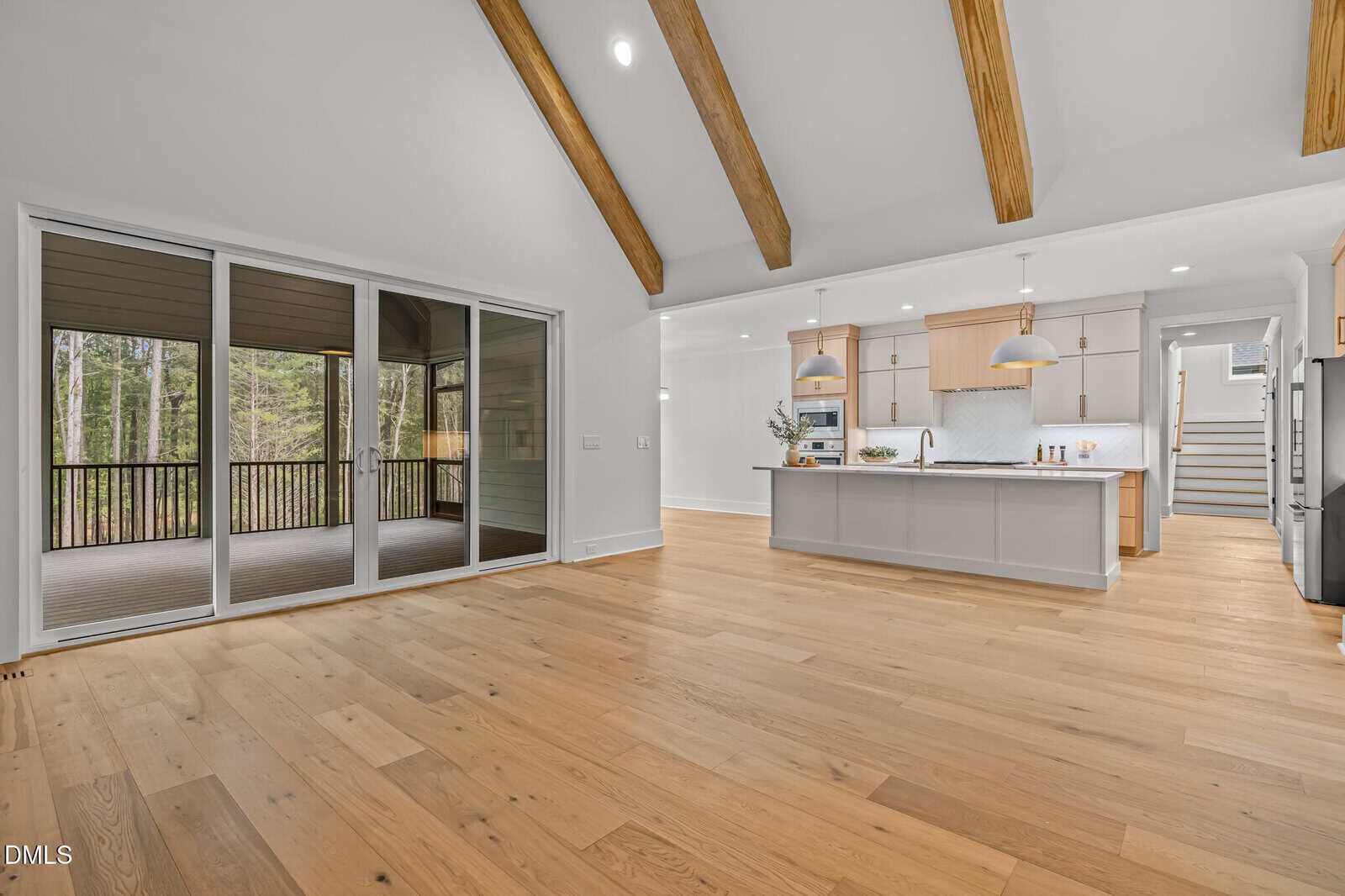 A spacious open-concept kitchen and living area with light wood floors, exposed wood ceiling beams, modern cabinetry, and large sliding glass doors leading to a screened-in porch overlooking trees.