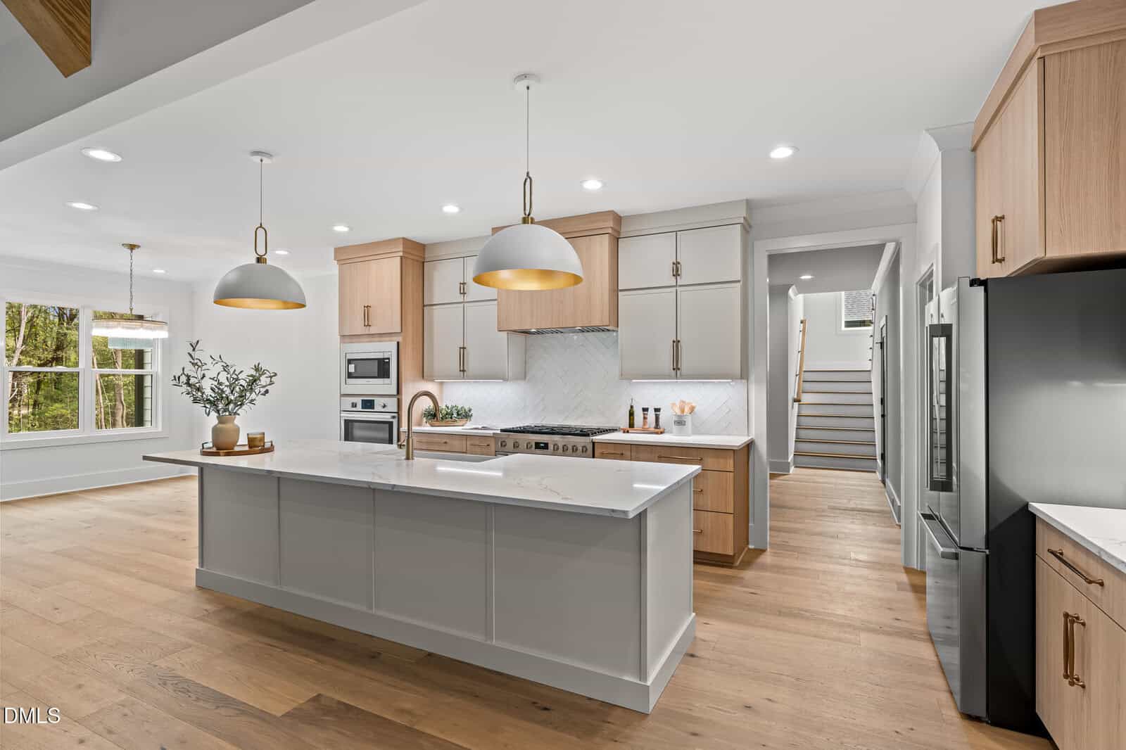 Modern kitchen with white and wood cabinetry, a large island with a built-in sink, pendant lights, stainless steel appliances, and light wood flooring. Stairs and a dining area with large windows are visible in the background.