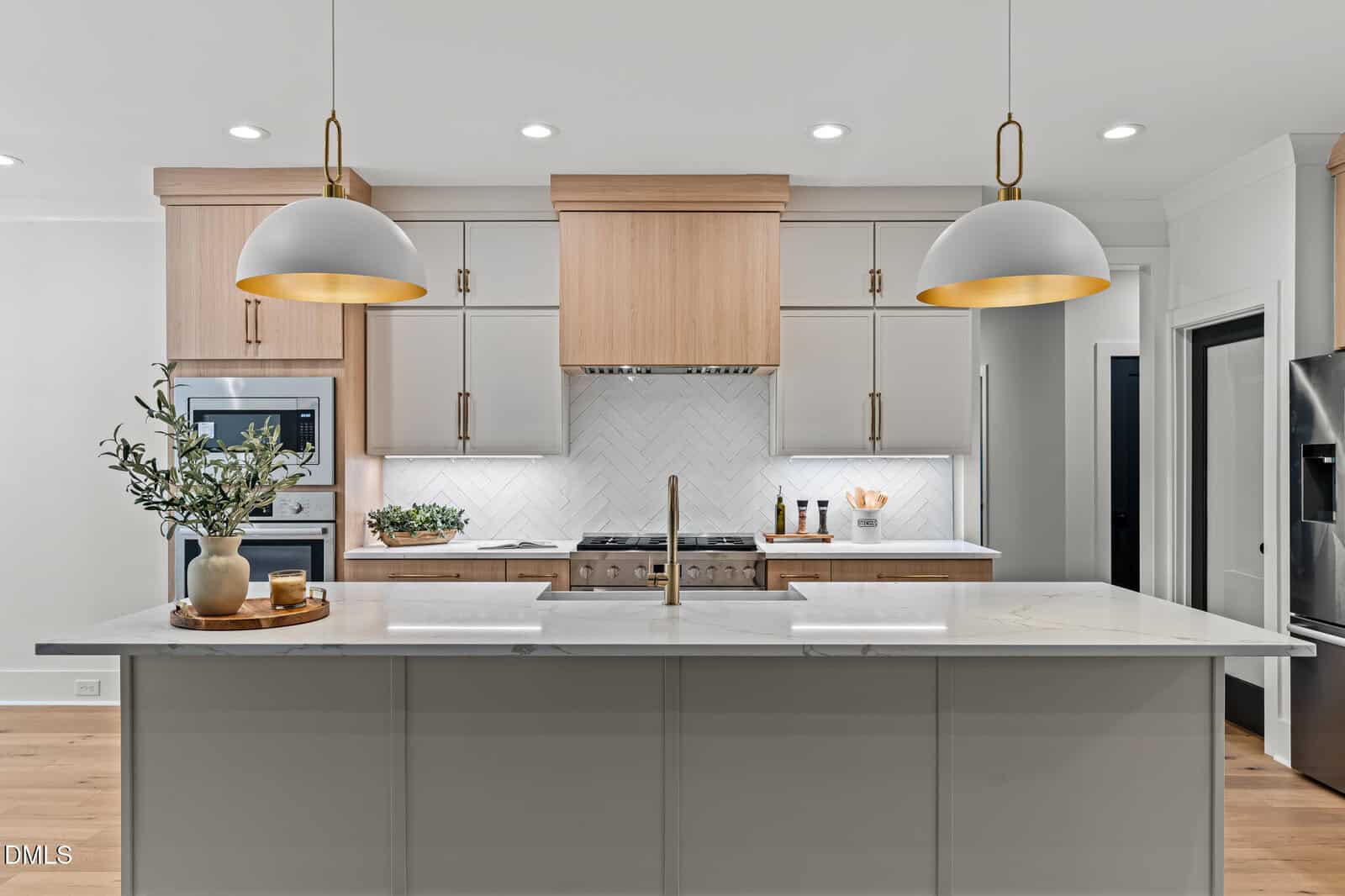 Modern kitchen with light wood cabinets, white countertops, two gold hanging pendant lights, stainless steel appliances, a central island with a sink, and decorative plants and items neatly arranged.