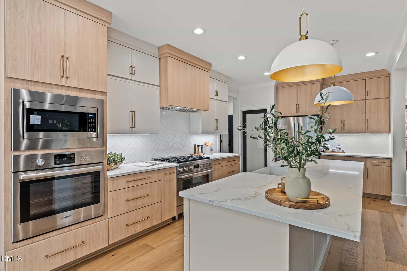 Modern kitchen with light wood cabinets, stainless steel appliances, a marble island with a vase of greenery, pendant lights, and herringbone backsplash. The space is bright, spacious, and neatly organized.