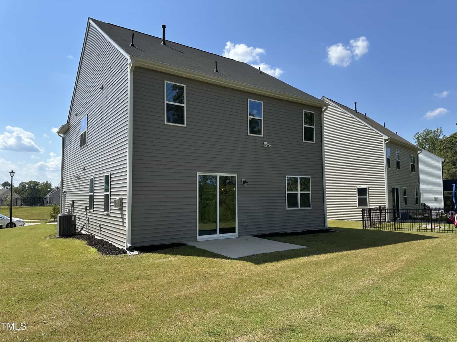 A two-story beige house with white trim and a black roof, featuring large windows and a sliding glass door leading to a small concrete patio in a grassy backyard on a sunny day.