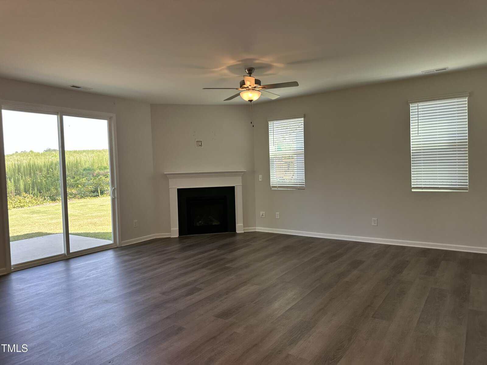 A spacious, empty living room with wood flooring, a ceiling fan, two windows, a fireplace, and a sliding glass door leading to a grassy outdoor area.