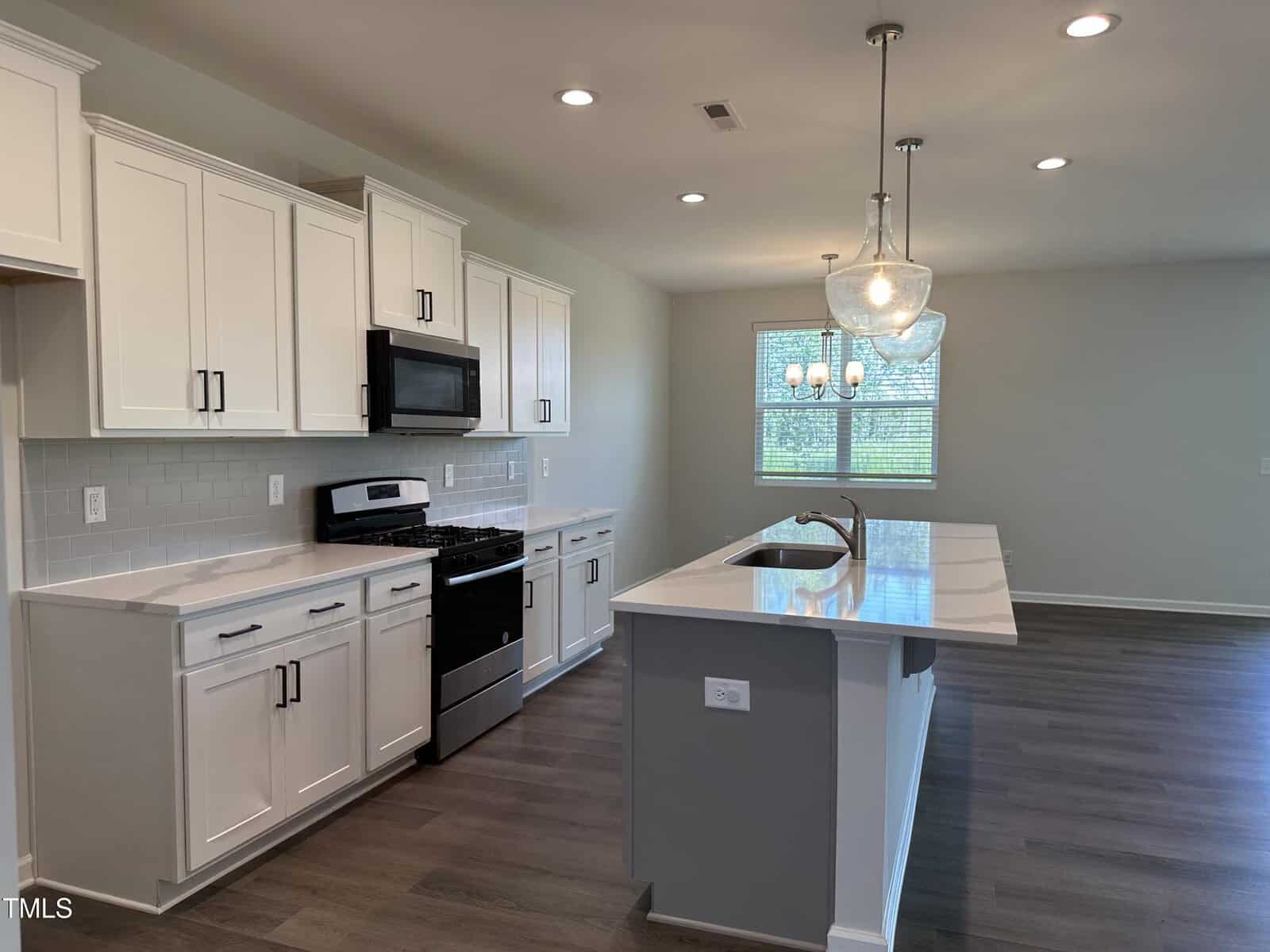 Modern kitchen with white cabinets, stainless steel appliances, a center island with sink, pendant lights, and wood-style flooring. Large window and dining area visible in the background.