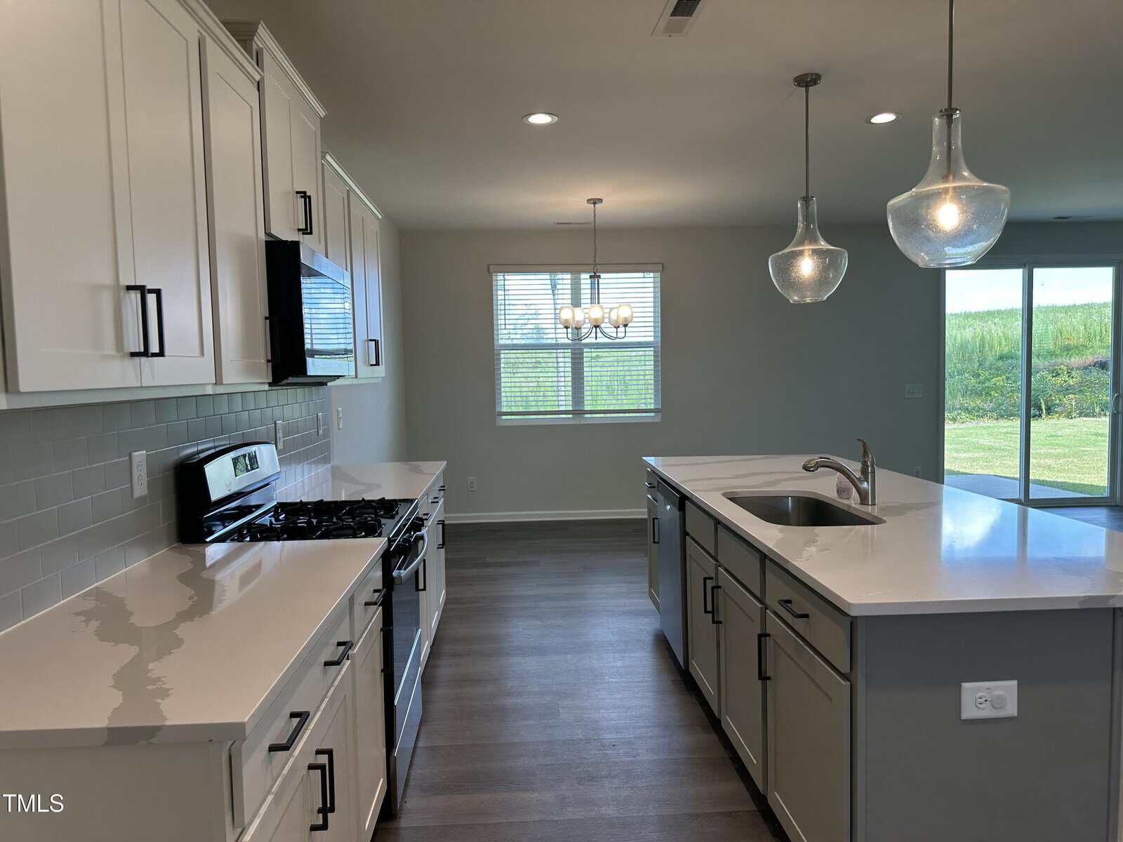 Modern kitchen with white cabinets, stainless steel appliances, a center island with sink, pendant lights, and large windows letting in natural light. Sliding glass doors open to a grassy yard.
