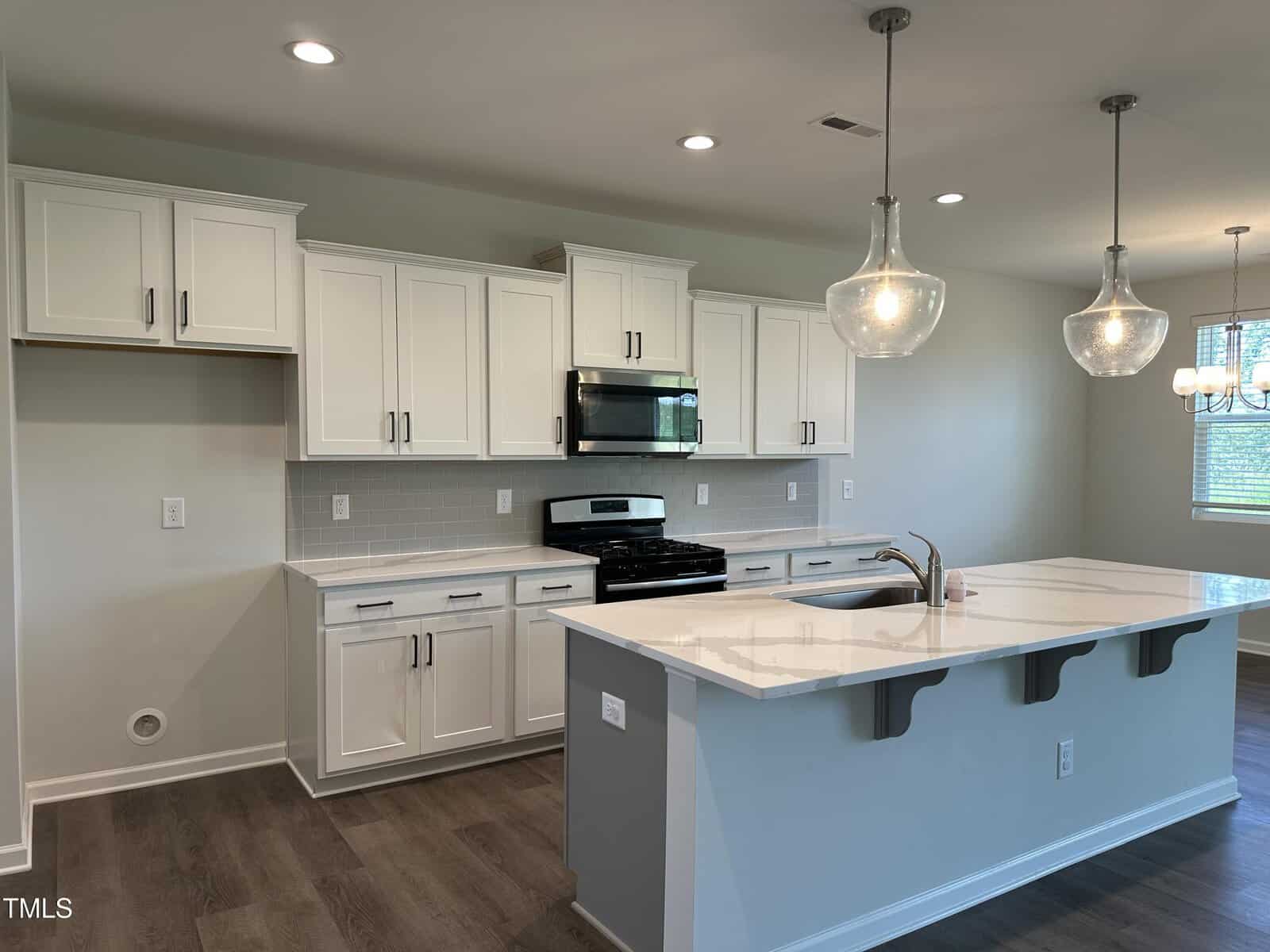 Modern kitchen with white cabinets, stainless steel appliances, a gas stove, over-the-range microwave, large island with sink, pendant lights, and dark wood flooring. Walls are light-colored, and windows let in natural light.