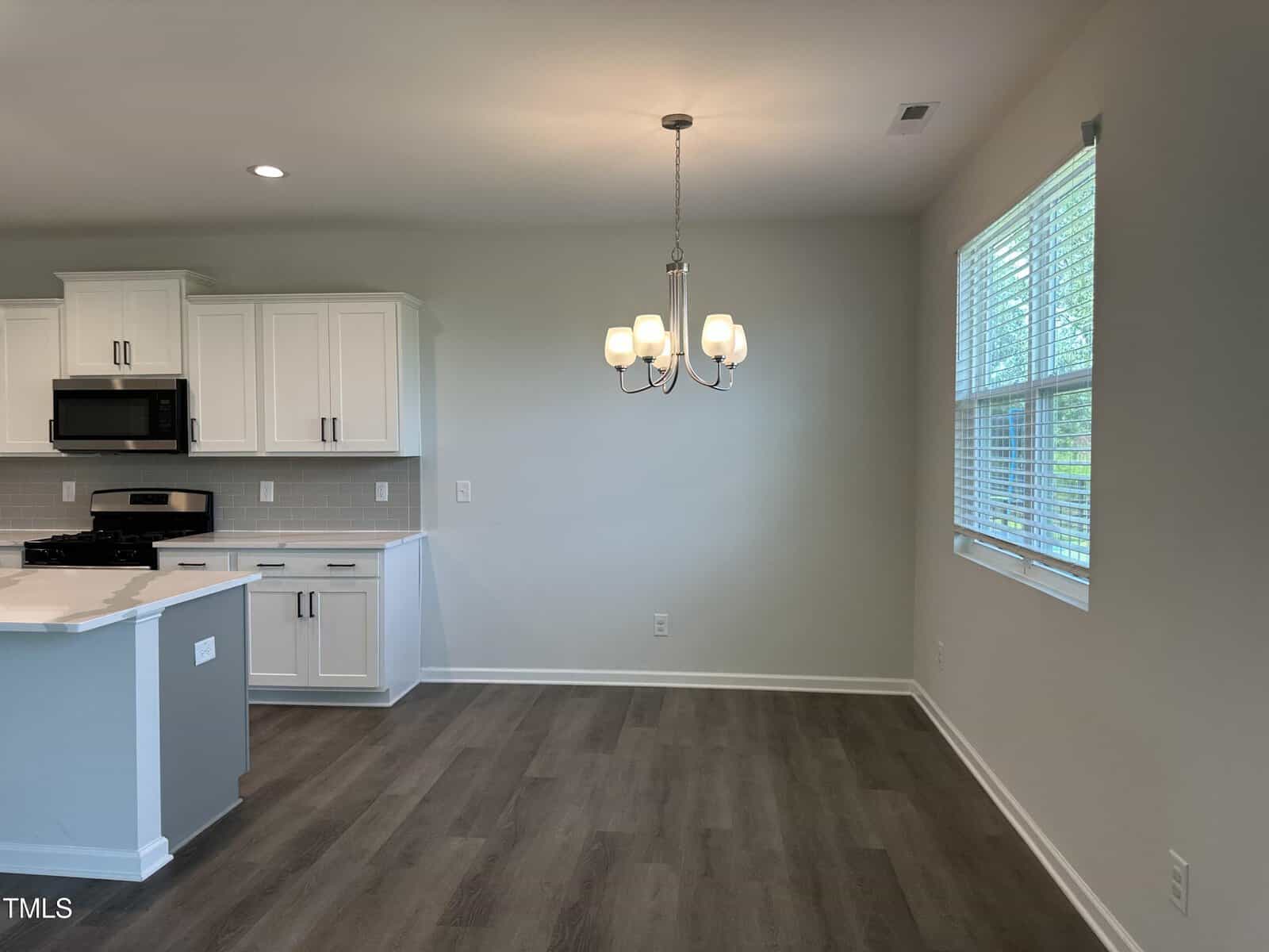 A modern kitchen and dining area with white cabinets, stainless steel appliances, a light fixture over the dining space, and large window with blinds. The room has wood-style flooring and light-colored walls.