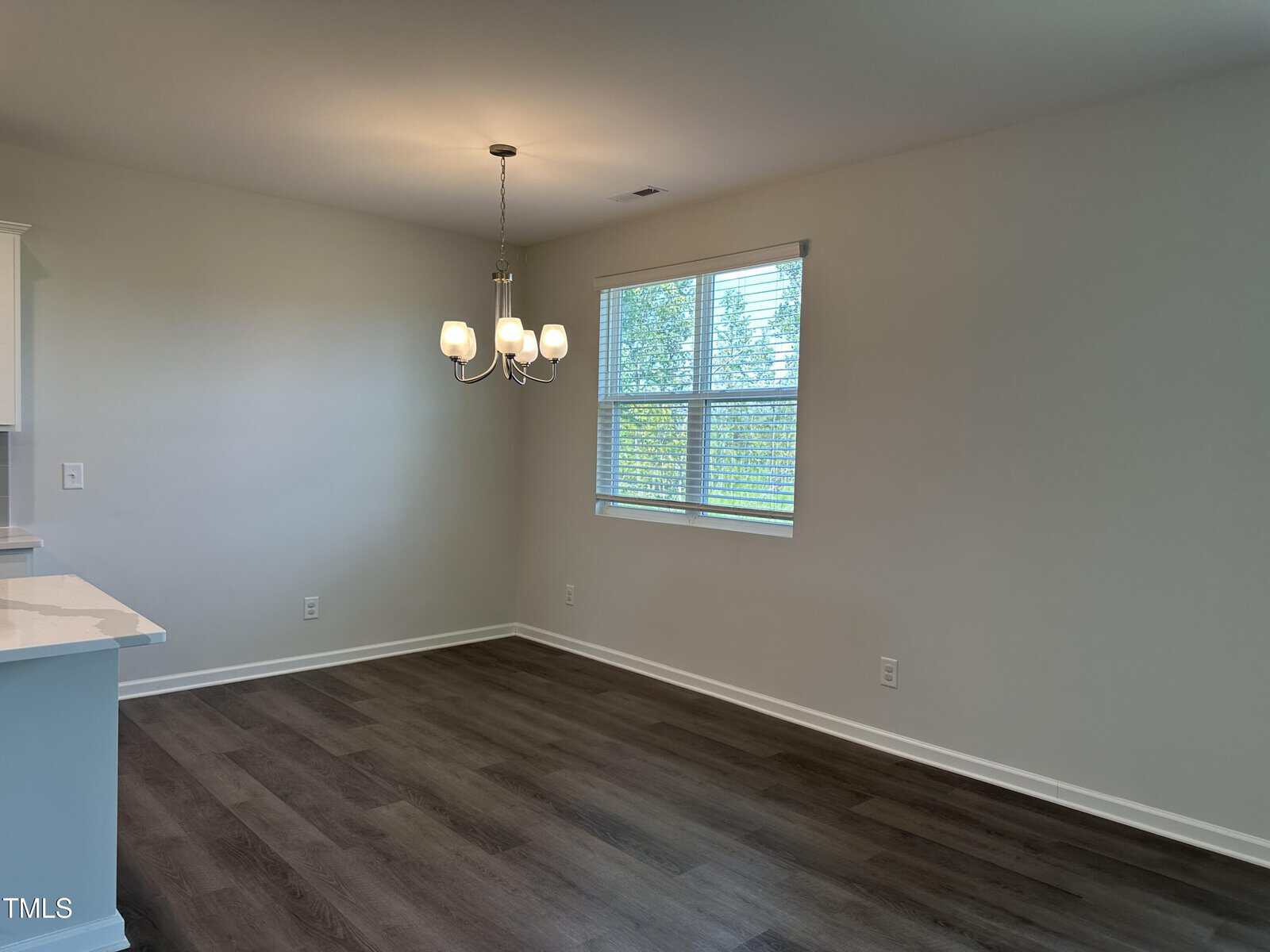 A dining area with dark wood flooring, a modern chandelier, and a window with blinds. The walls are light-colored, and sunlight comes through the window, creating a bright, clean atmosphere.