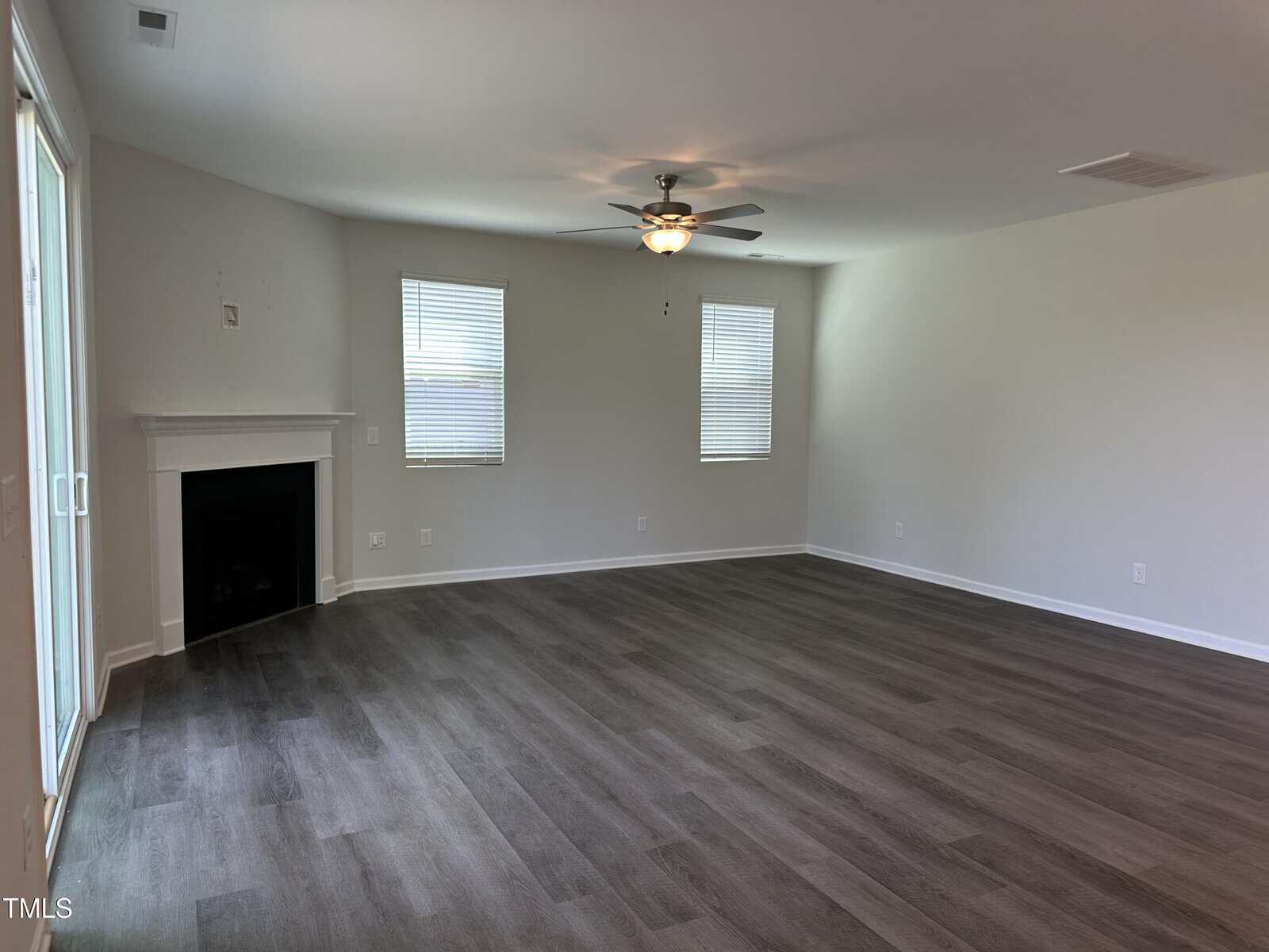 Spacious empty living room with light gray walls, two windows, a ceiling fan, a corner fireplace, and dark wood flooring. Sliding glass doors are visible to the left.