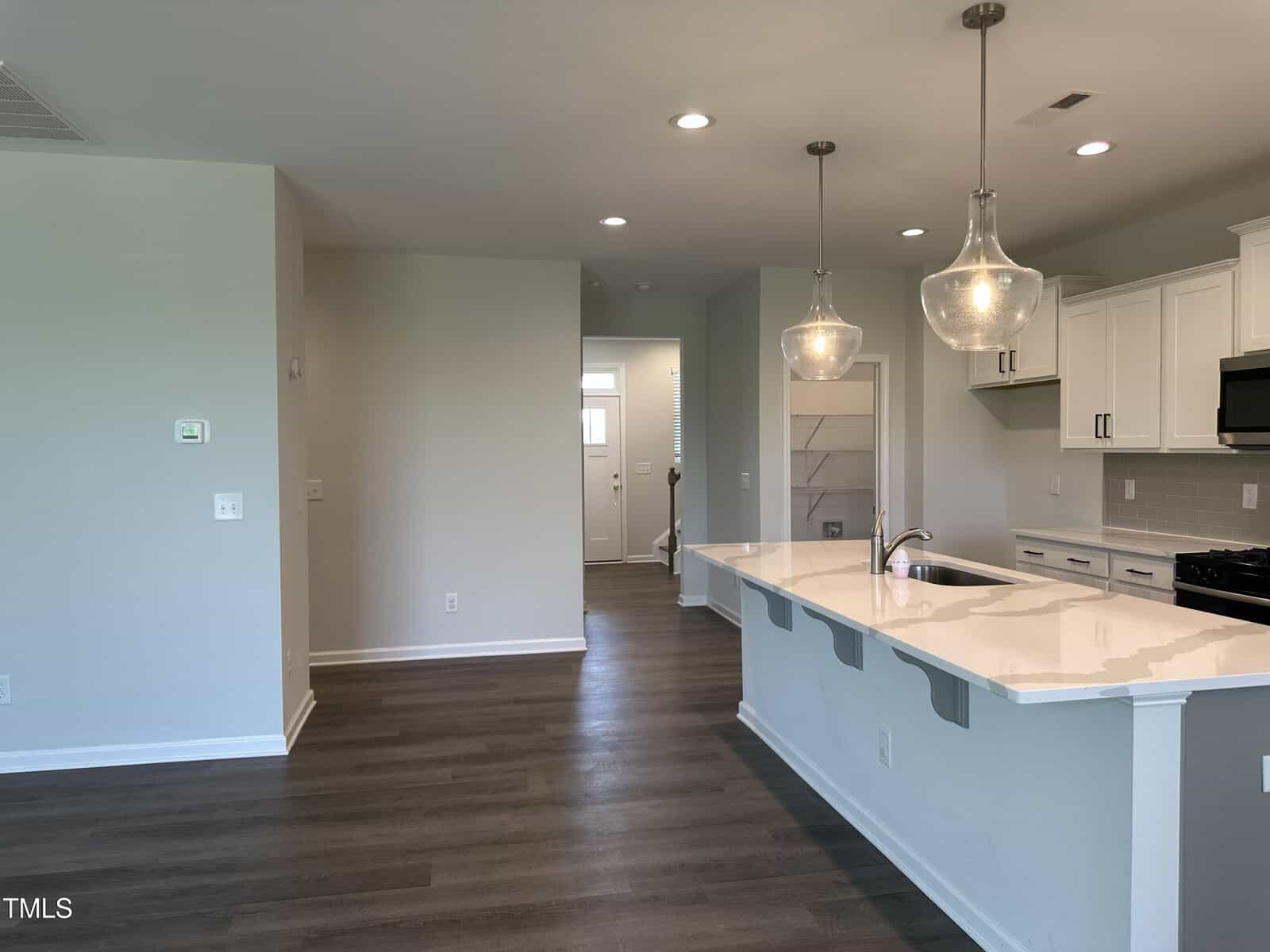 Modern kitchen with white cabinets, quartz countertops, two pendant lights, and stainless steel appliances. The area has wood-look flooring and a view into a hallway with natural light from a distant door.